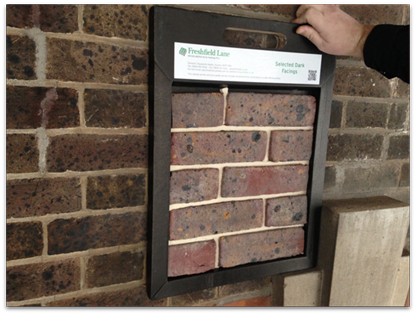 Image is of a brick wall with brown bricks. A sample panel of brown bricks is being held up to see if it matches {{brizy_dc_image_alt imageSrc=