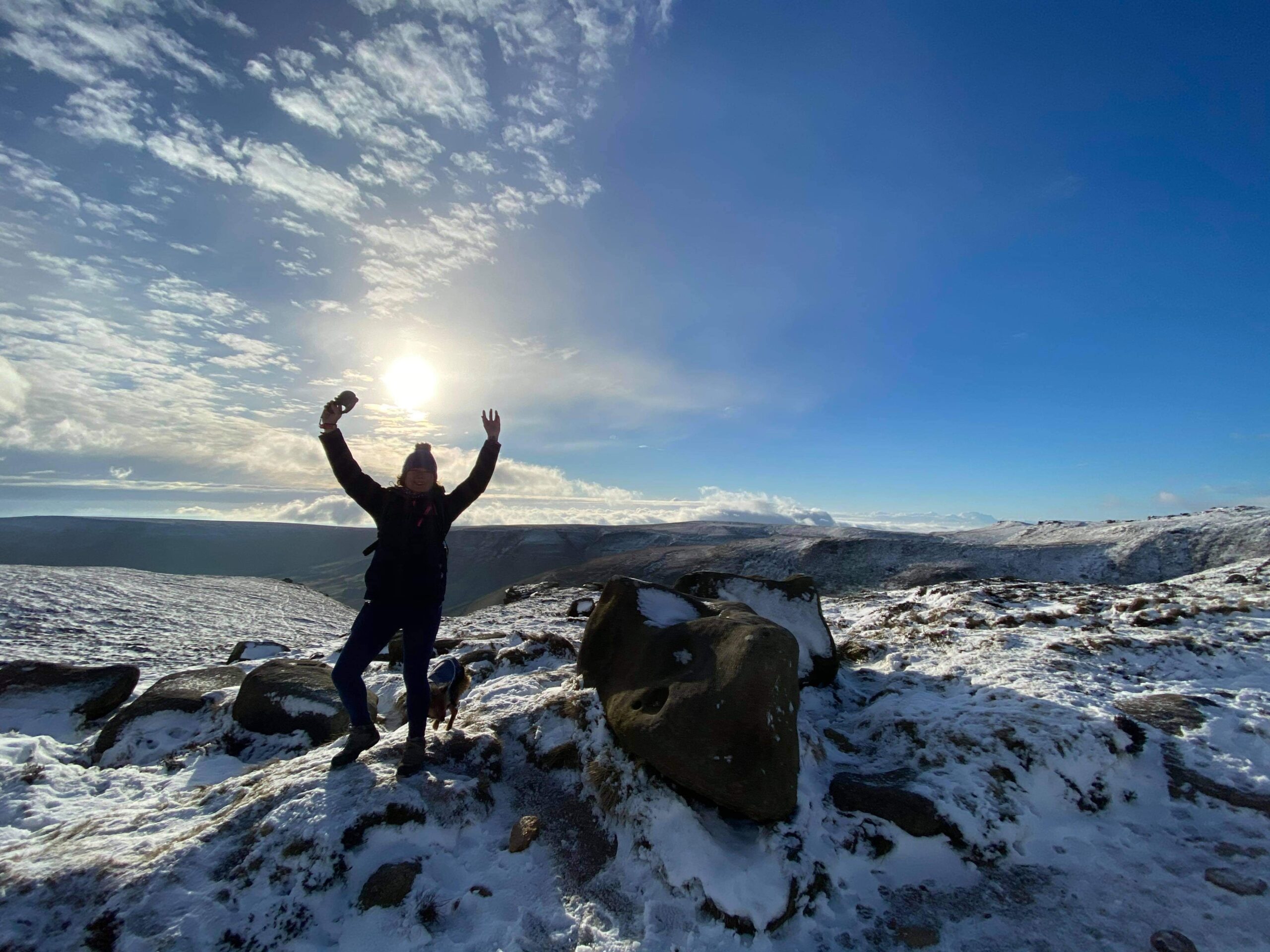 Silhouette of Ruth on a sunny winter's day, with her arms in the air on top of Kinder Scout Mountain in Derbyshire, with some snow on the ground.