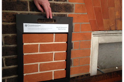 Image is of a brick wall with brown and red bricks. A sample panel of the red bricks is being held up to see if it matches
